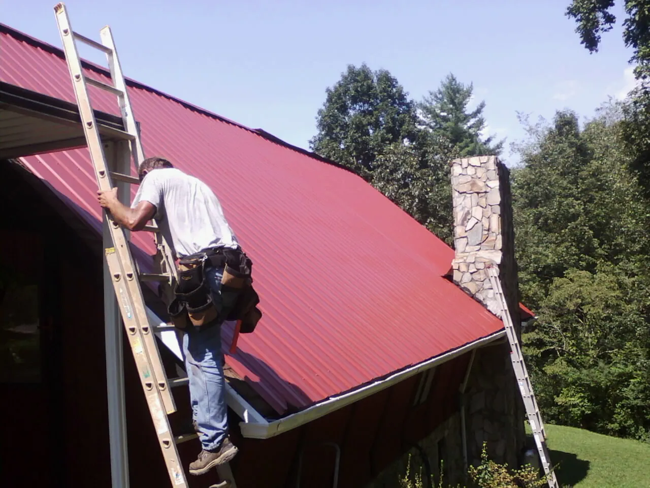 Roofer on a ladder beside a red metal roof and stone chimney — Soffit & Fascia Repair by Roofing Brothers in King Ferry